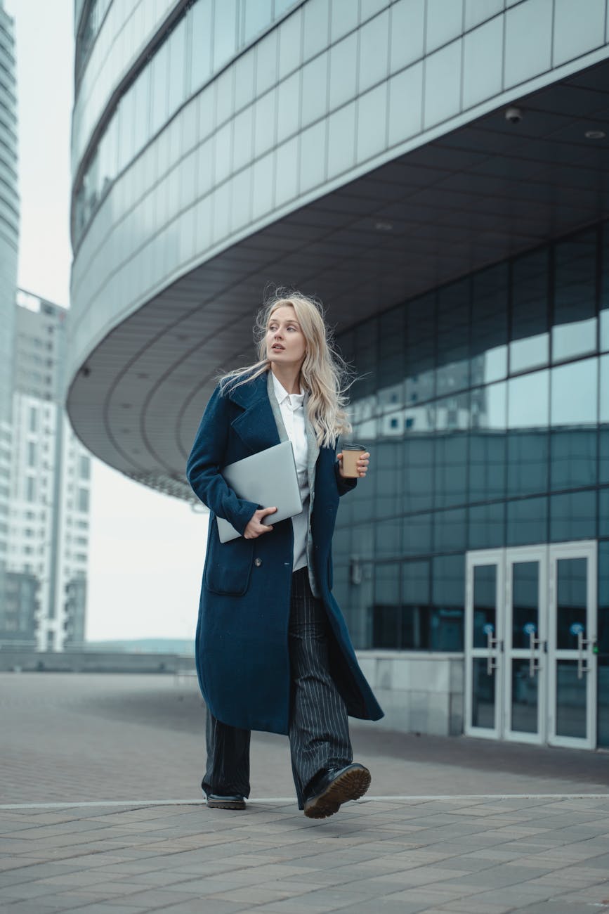 Modern businesswoman with laptop and coffee walking in urban setting.