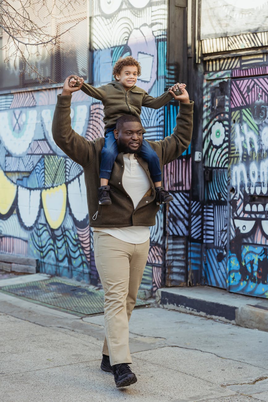 Full body of positive African American father with boy on shoulders strolling on sidewalk near wall with graffiti in city