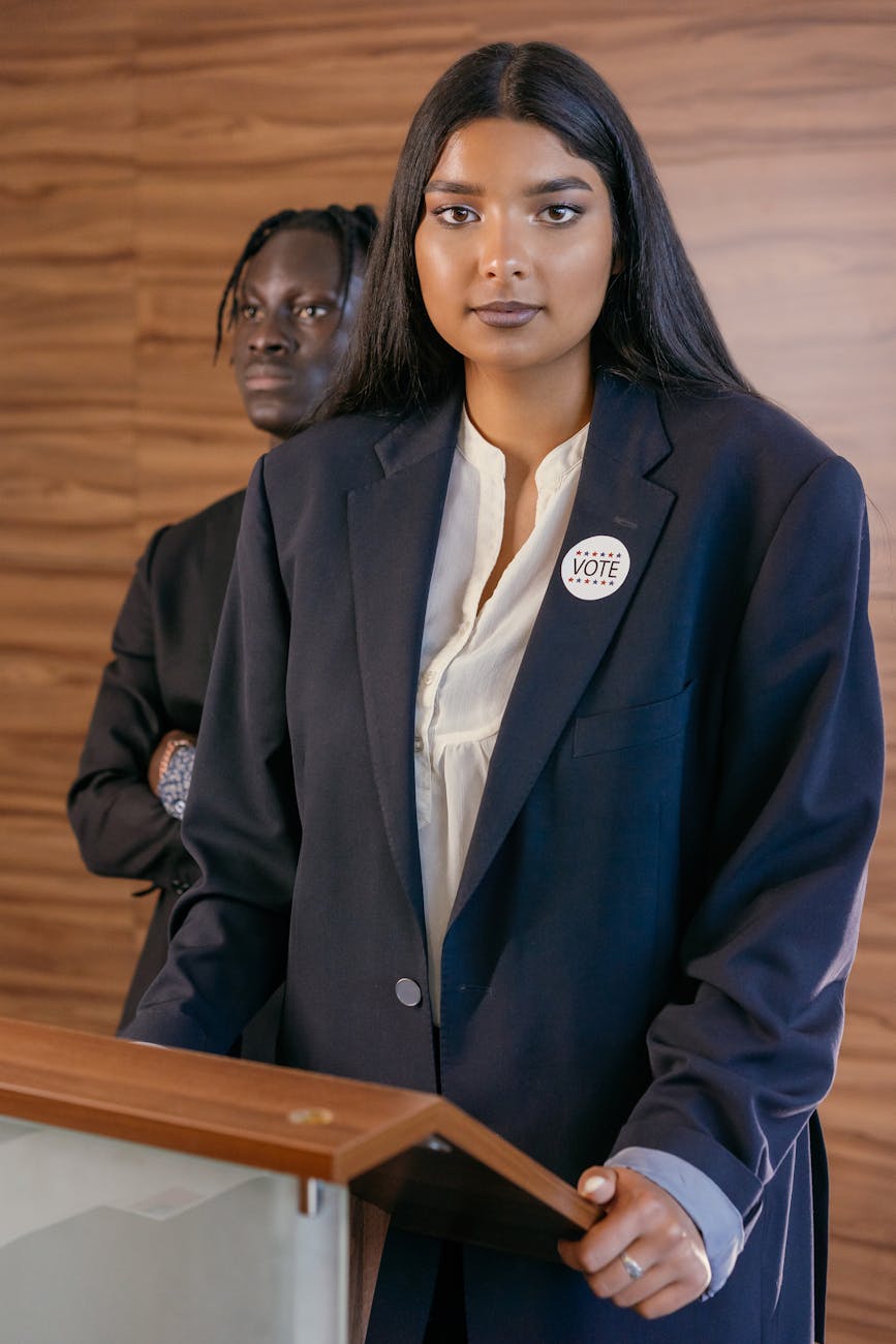 A woman in a blazer stands at a podium with a vote badge, promoting leadership and participation.
